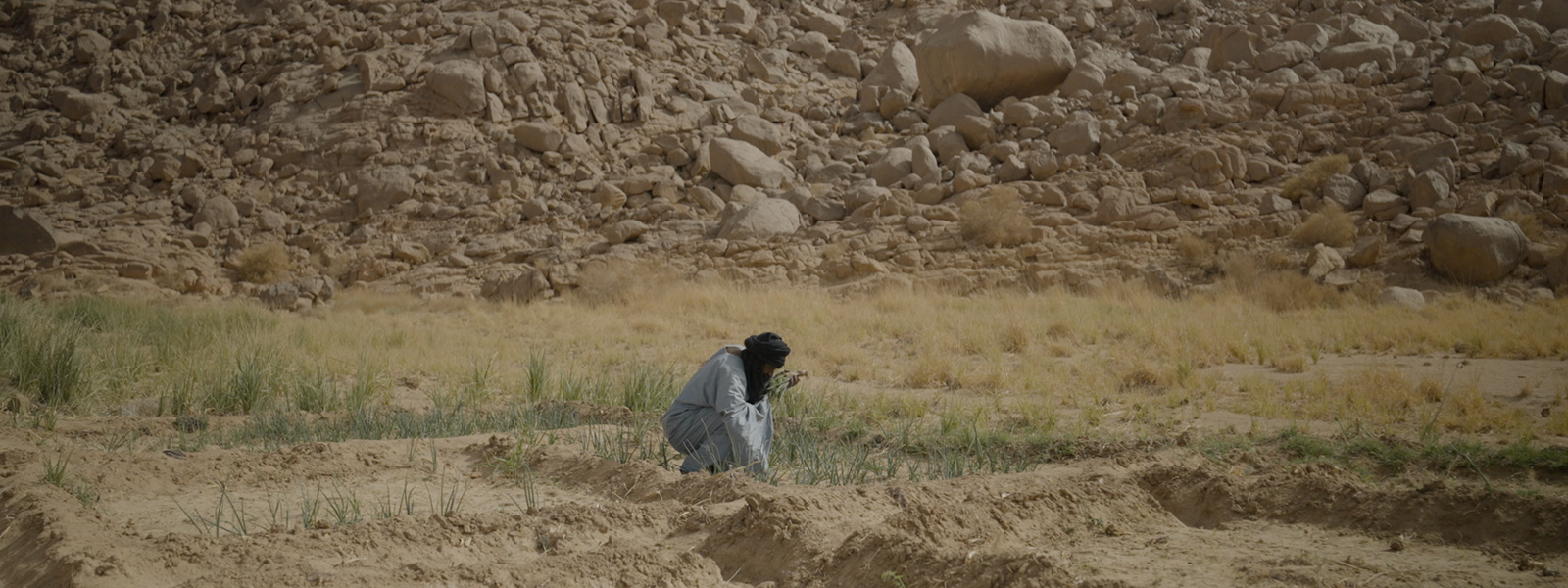 A person in light clothing and a dark headscarf crouches down in the desert looking at the ground