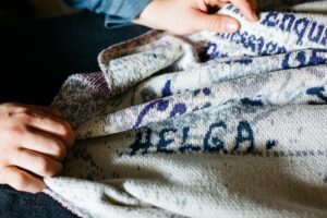 Two hands touch a knitted blanket. The blanket is cream with blue woven text. The only word that can be made out is 'HELGA'.