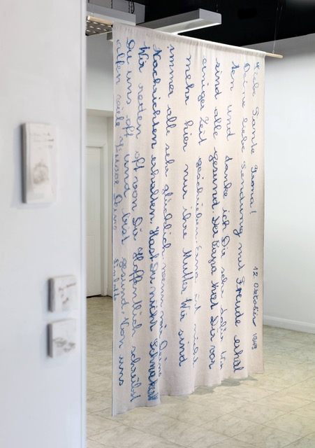 A large blanket hangs in a gallery, suspended from the top edge. The knitted blanket is off-white, with blue knitted text. The piece looks like a letter or handwritten note.