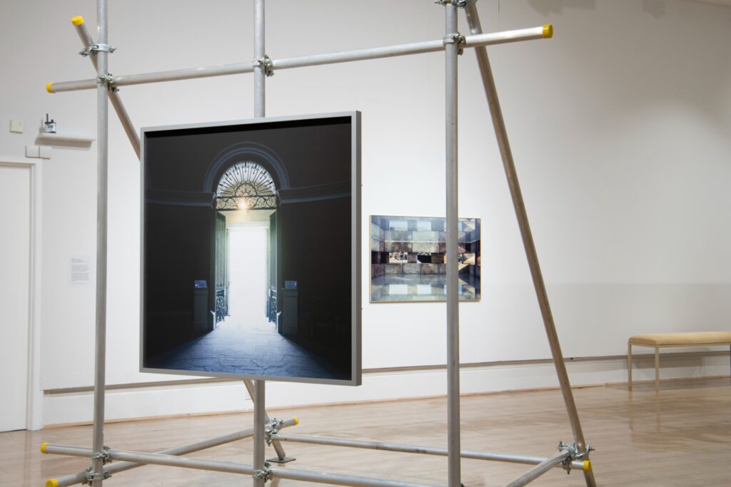 The Sunderland Museum gallery space. In the foreground a large colour photograph of a doorway in a square frame is supported by a frame of scaffolding. Behind on the white gallery wall a colour photograph of a scene inside a box.