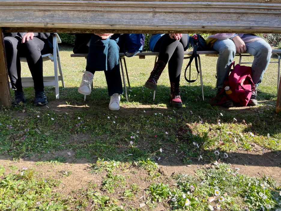 A shot of four sets of legs in jeans and leggings under a table top in a grassy field, taken during an outdoor seminar.