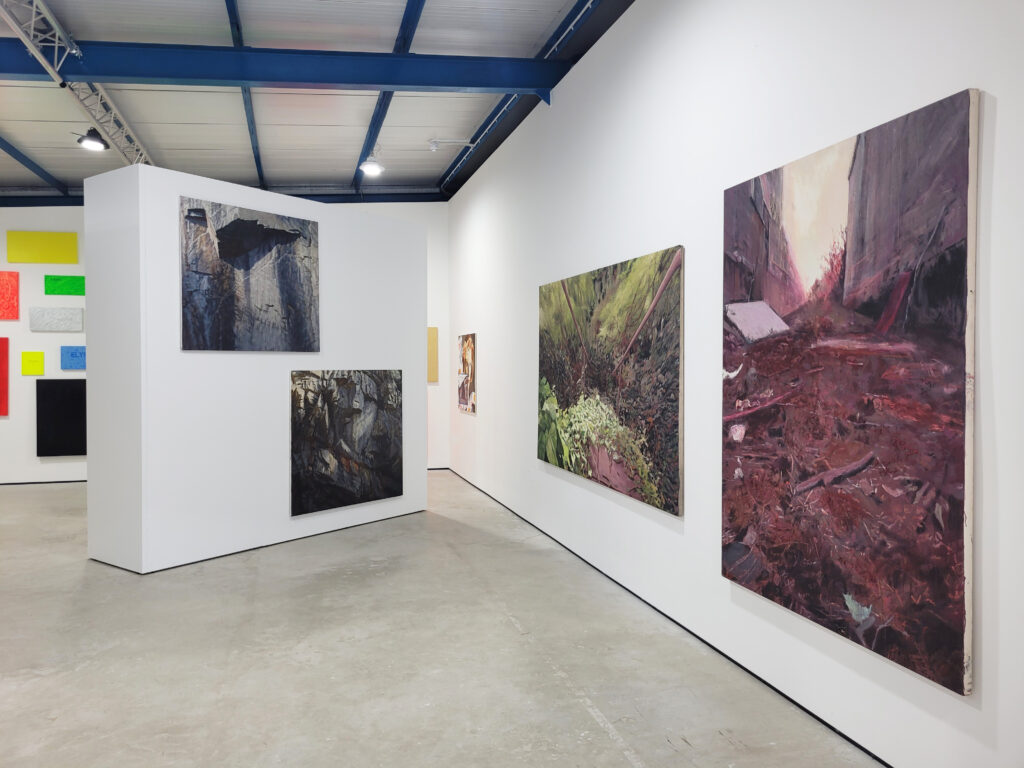 Installation view that includes four large square canvases with dark coloured paintings on them in greens and browns and blues