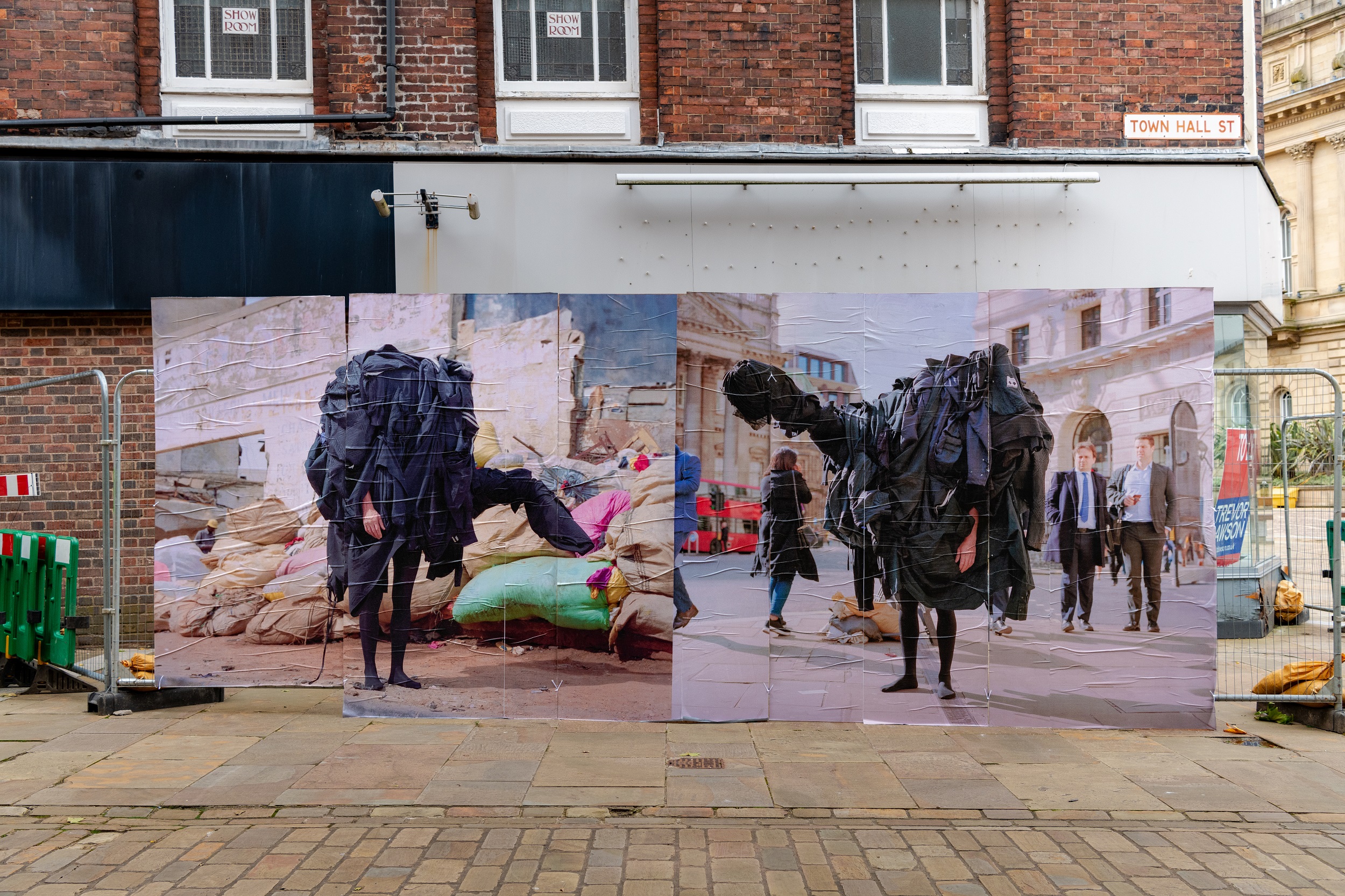 A construction hoarding on a town centre street pasted with two images from Hutchison's project, showing his zombie sculpture outfit made of black clothes, on the left in front of huge backs presumably full of second hand textiles and on the right in central London.