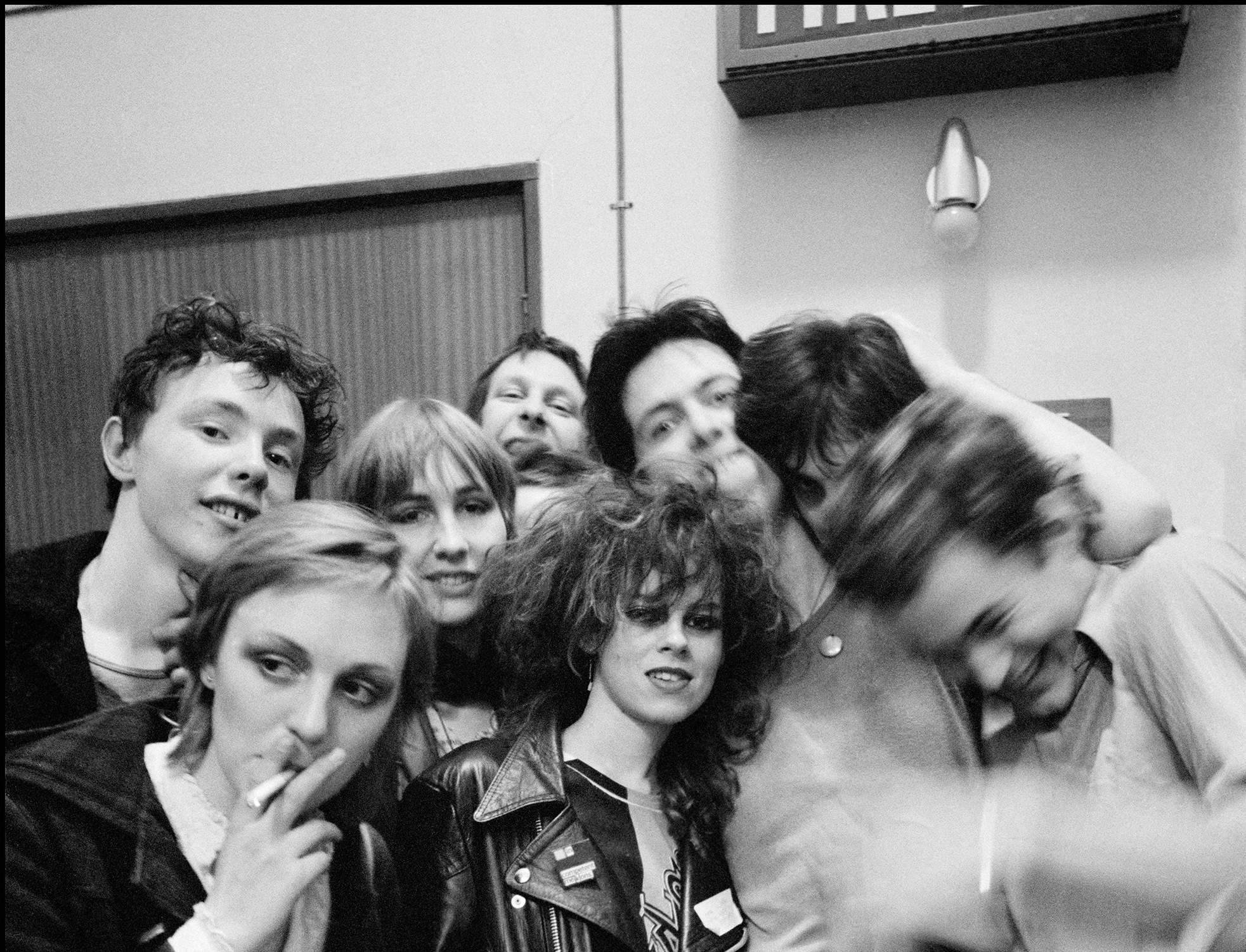 A black and white photograph of Mekons, a group of young white people in 1979 justling with each other, drinking and smoking