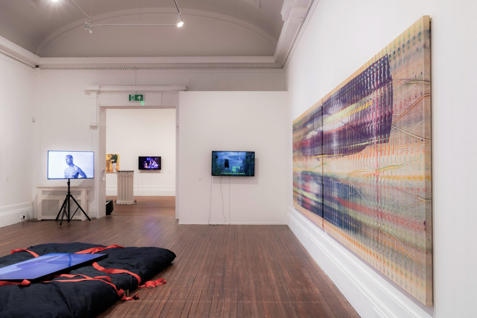 The gallery has white walls and brown wooden floors. A large multicoloured canvas to the right with small monitors against the far back wall and a large black cushion on the floor in the middle of the space.