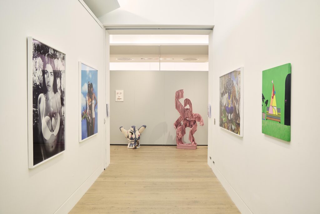 View down a corridor, two paintings on each of the side walls, at the far end, three sculptural forms against a grey wall - a ceramic face, a knitted woollen torsa, a figure made from draped pink fabric on an aluminium frame.