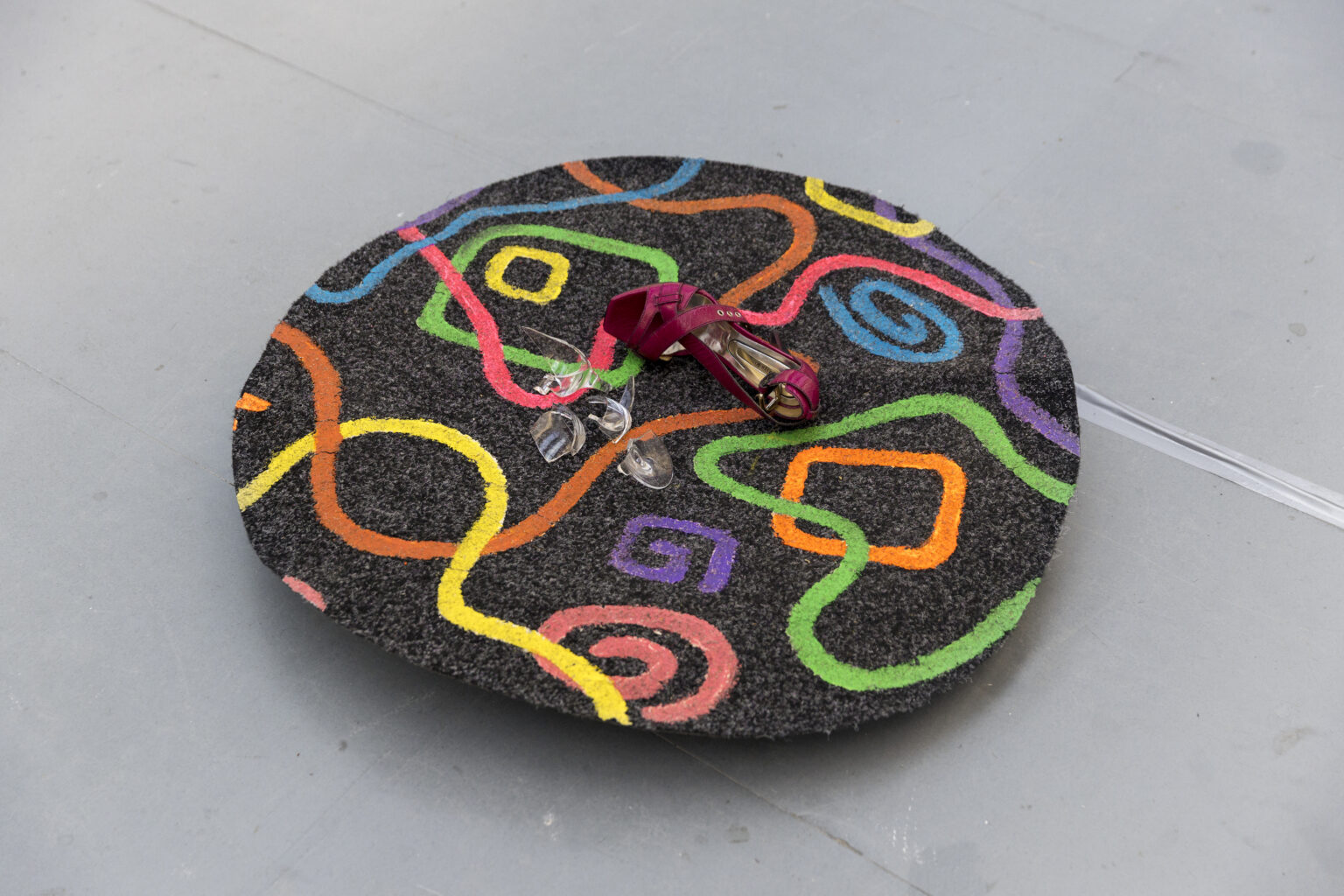 A disc about a meter in diameter, cut from what looks like a dark grey carpet tile is set a few inches from the ground. The disc has brightly coloured squiggles and shapes painted on to it. there is a broken glass and a pink high heeled shoe laying on the top.