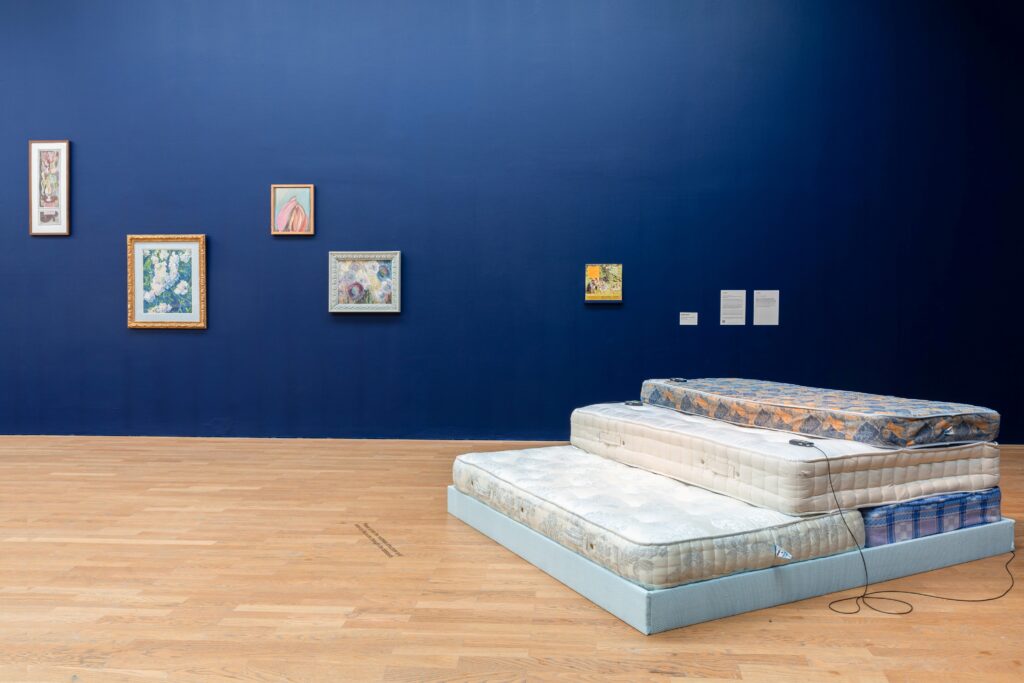 Dark blue wall with framed paintings, stack of mattresses in the foreground.