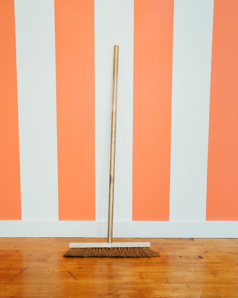 A broom with a bronze handle rests against an orange and white striped wall