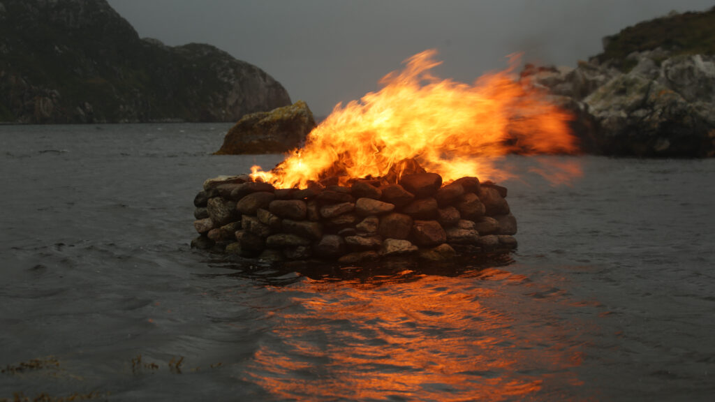 A pile of stones stacked up our of the surrounding sea at night, with orange fire on top blowing in the wind towards the right of the frame