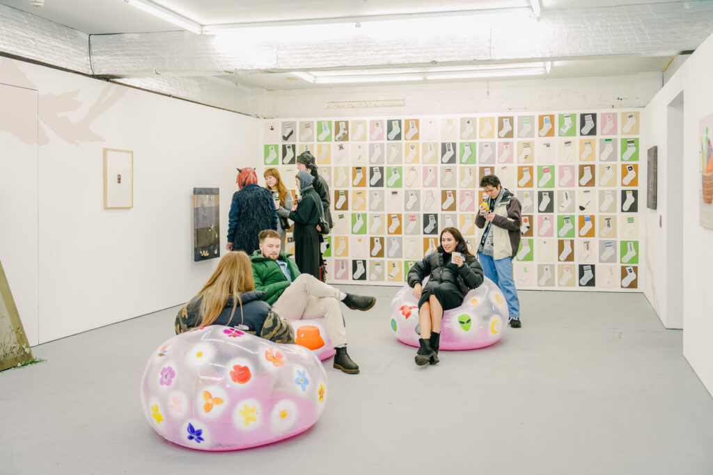 A white walled gallery space with drawings of socks on colourful backgrounds covering one wall, and people sitting in the foreground on inflatable chairs.