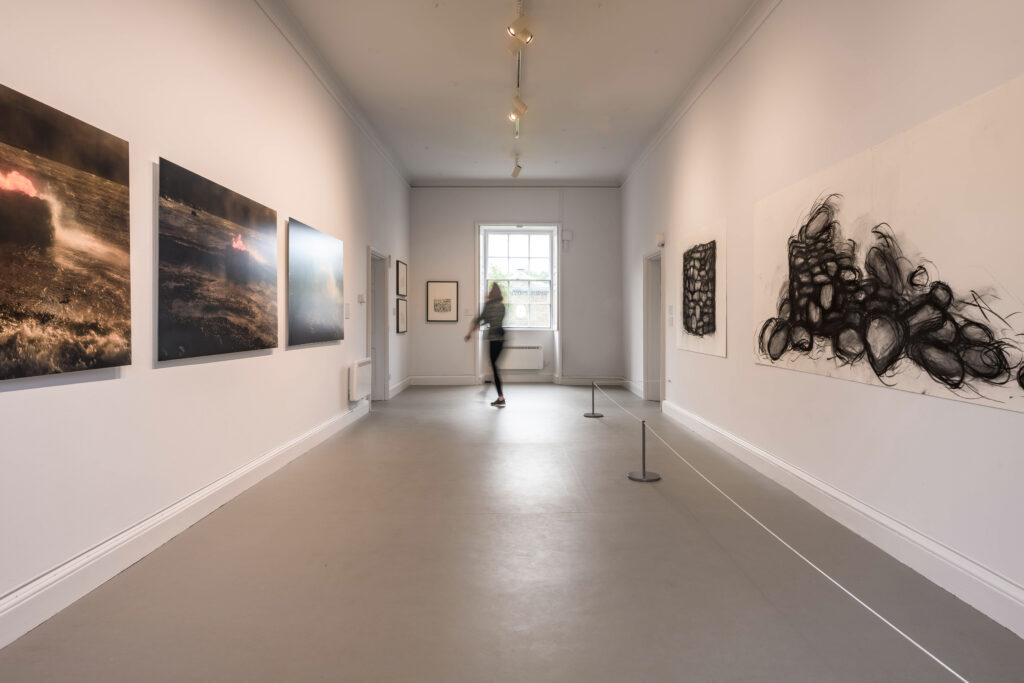 A white wall gallery space with printed photos of natural formations in dark colours on the left and large charcoal drawings on white of piles of stones on the right