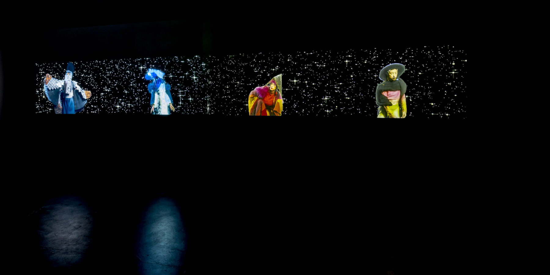 Four screens in a dark room show characters in brightly coloured costumes against a starry sky