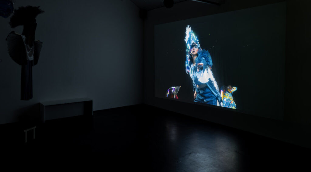 A darkened room: to the right, a screen showing a character in a brightly coloured costume against a starry backdrop; to the left, a costume hanging in the darkness.