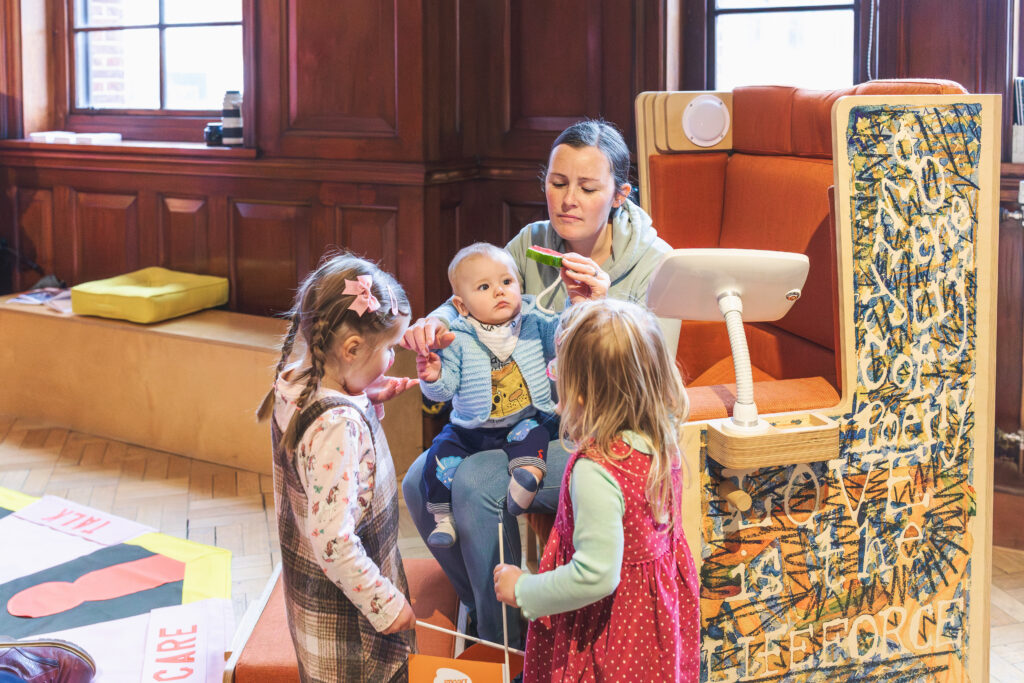 A woman with a baby and two older children sit in the Feeding Chair, which is a decorated wooden structure, within a wood-panelled gallery at The Tetley.
