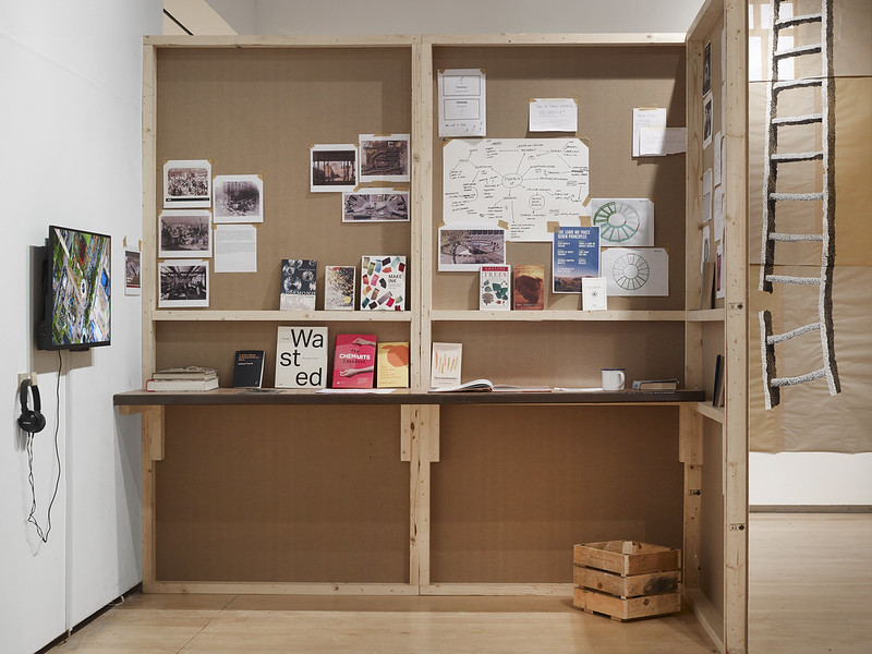 Installation view of the replica studio space built of plywood with research, various printouts and diagrams on the walls and books propped up on the bench. a TV screen on the wall to the left with headphones