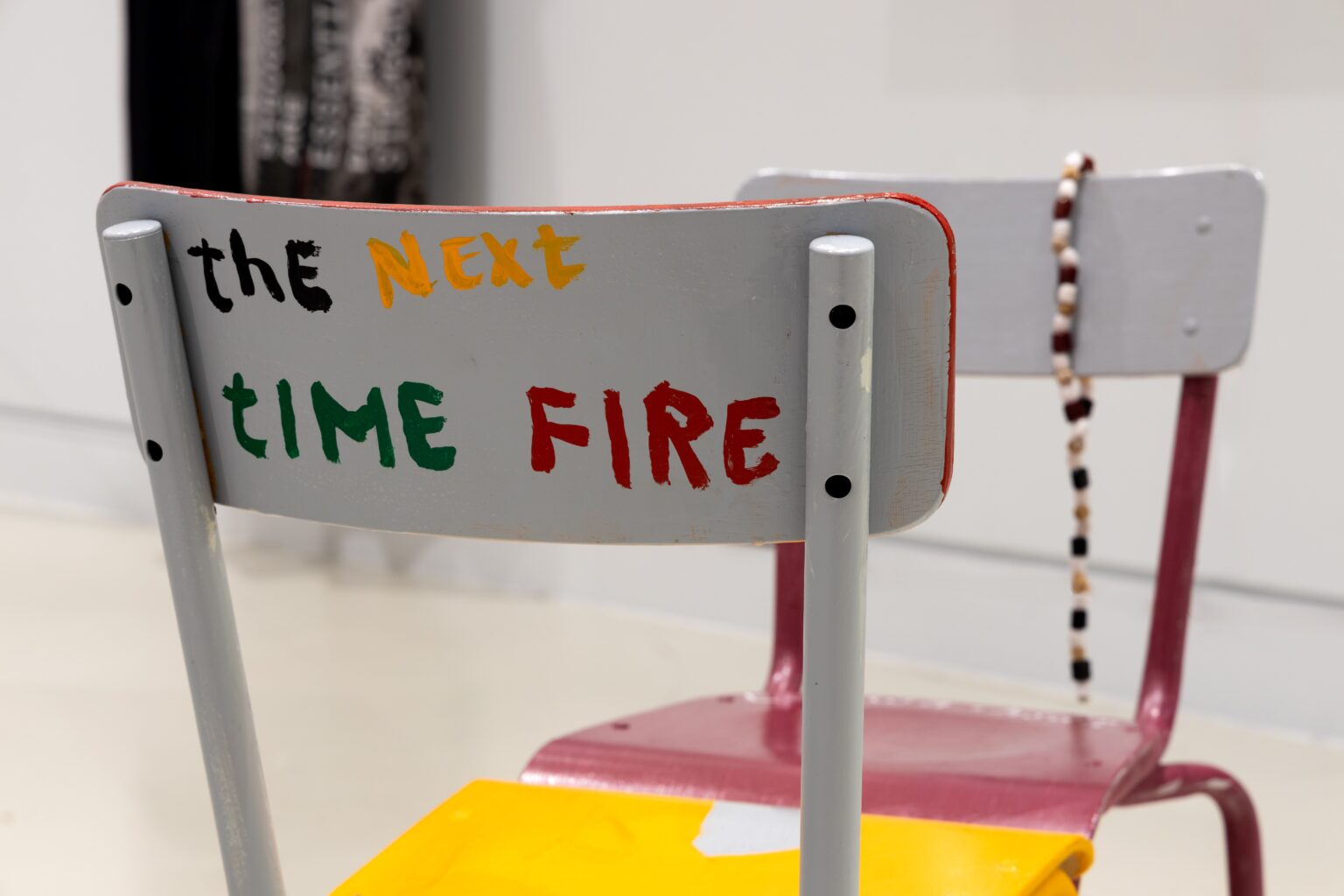 Two painted school chairs, lips of seats touching. Beads hang from the backrest of one, the other has the words 'thE Next tIME FIRE' painted on the back.