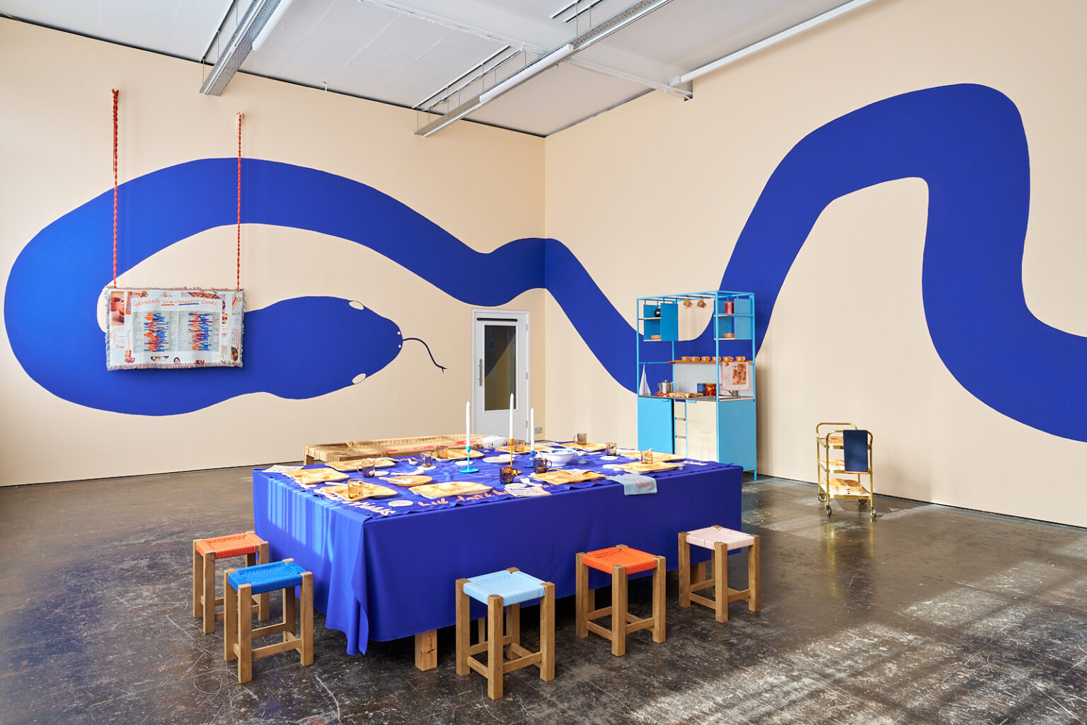 An installation view of Courses for Dis-Courses by Roo Dhissou showing a laid table with a royal blue tablecloth and stools in different colours, the walls are painted pale peach with a mural of a snake in the same royal blue as the tablecloth curling around the room.