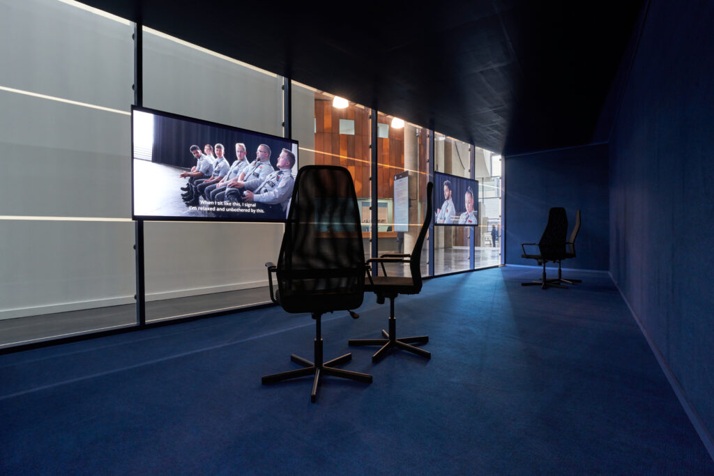 Four computer chairs are gathered around two TV screens showing indistinct seated figures. The screens are mounted on one-way glass, through which you can see FACTs foyer.