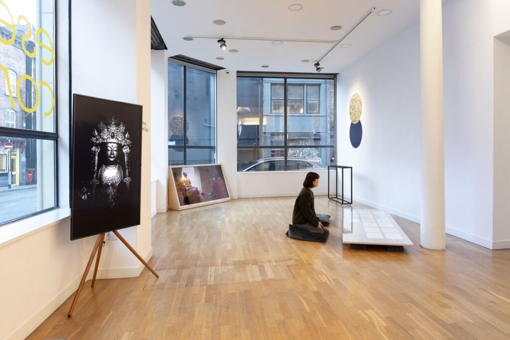 A bright white gallery space with a viewer sitting on a floor cushion in the centre of the room looking into a low vitrine