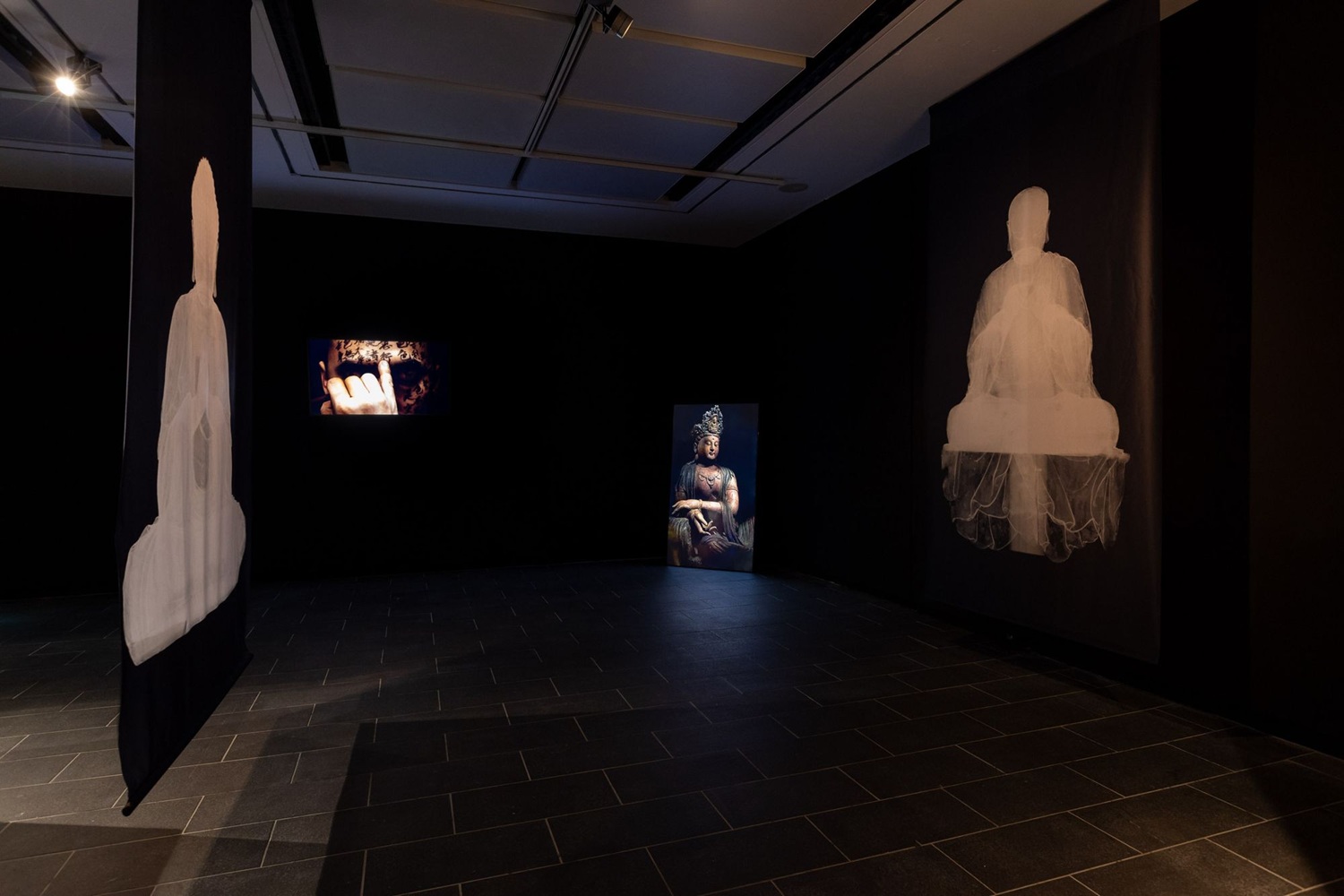 A dark gallery space with two prints of Buddha statues in white and two screens, one showing a low lit statue and on the other an image of a human hand