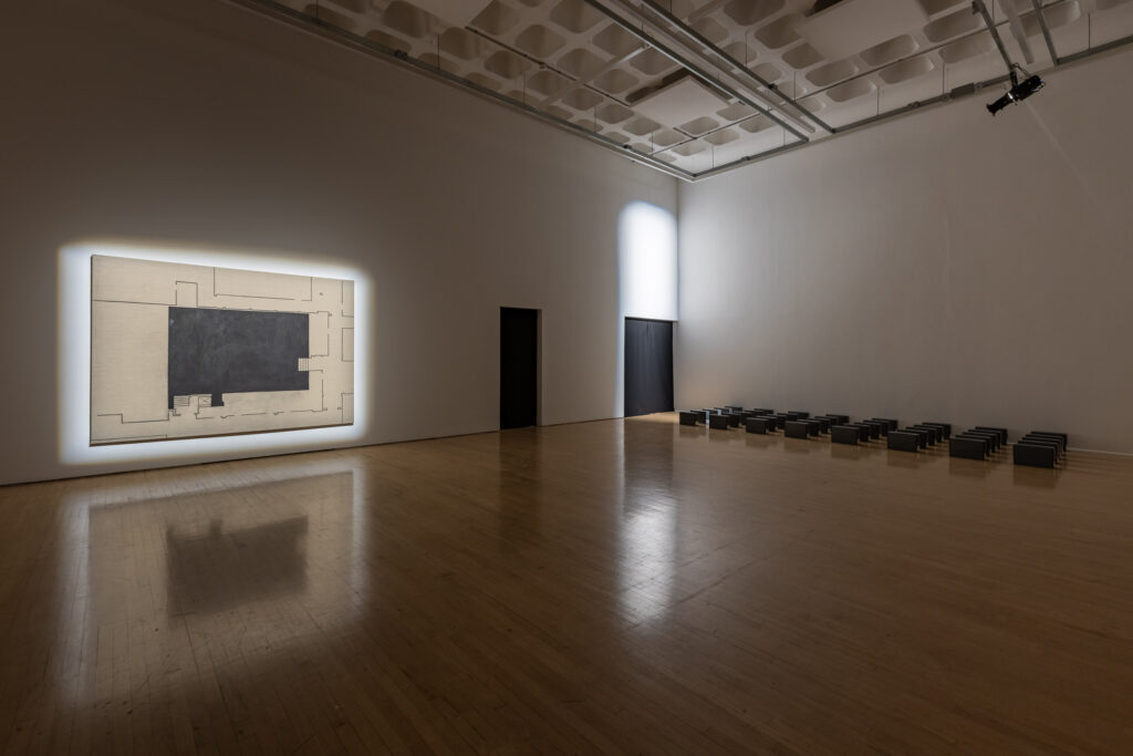 A darkened, white-walled gallery space with concrete roof and wooden floor. Spotlights pick out a large canvas with charcoal drawing and a black-curtained exit on the back left-hand wall, with rows of dark-grey archive boxes lined up as if about to leave through the exit.