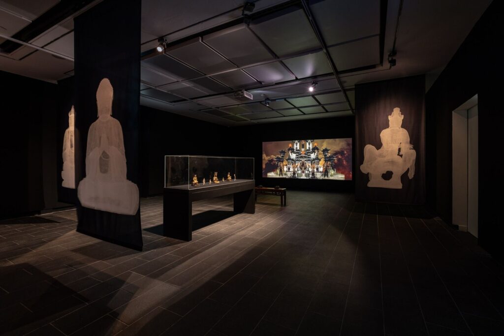 A dark gallery space with three lit up white images of Buddha statues, a vitrine in the centre with small golden figurines, and a large screen on the back wall with a temple