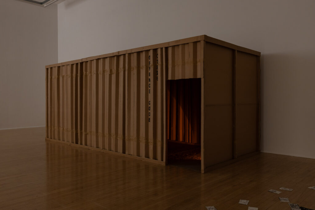 A shipping container made from cardboard sitting on a wooden floor in a white-walled gallery space. There is a door cut in one corner of the container, at the edge of one of the longest sides.