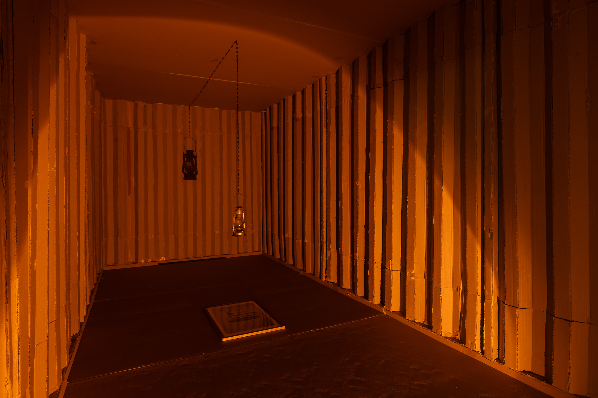 The interior of a shipping container made from cardboard. A yellow-orange light picks out an oil lamp hanging from the centre of the roof, a framed image lying flat of the floor beneath it, and the ridged interior walls of the shipping container.