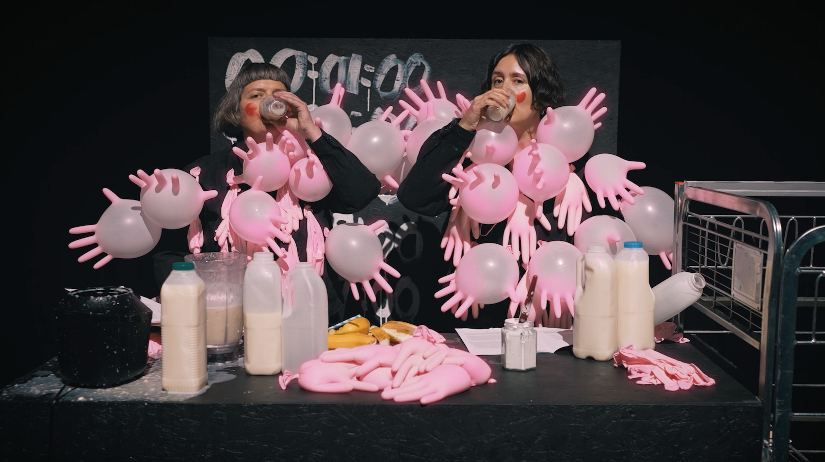 Two women artists wearing black black and covered in pink blown-up surgical gloves drink milk from glasses, looking at the camera and surrrounded by pints of milk.