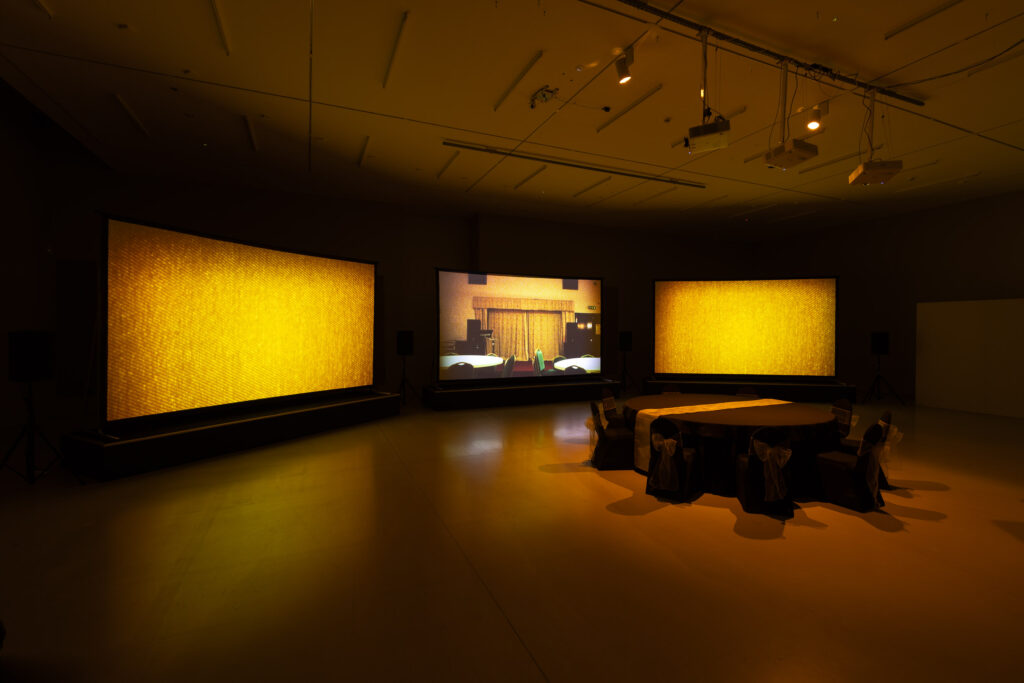 A darkened gallery space with three projection screens arranged before a table and chairs arranged as if for a special event. The screens on either side show yellow fabric in close detail, and the central one shows a stage with curtains of a similar colour.