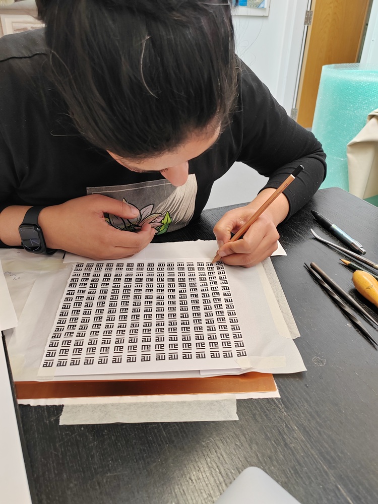 the artist is bend over a sheet of paper drawing rows and rows of the word 'me'