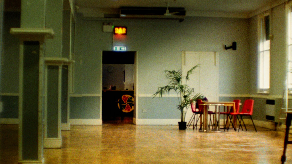 An empty community space with parquet flooring and sage-green walls. A table in the back corner and a darkened doorway leading through to the bar.