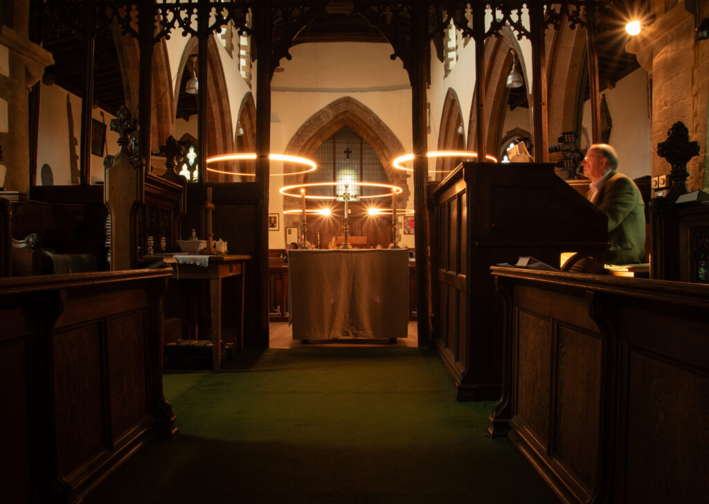 Five lights trace a circular path as they rotate, suspended from the ceiling of a church and captured on a slow shutter speed to show the arc of their movement. The lights glow warmly and illuminate the faces of the congregation.