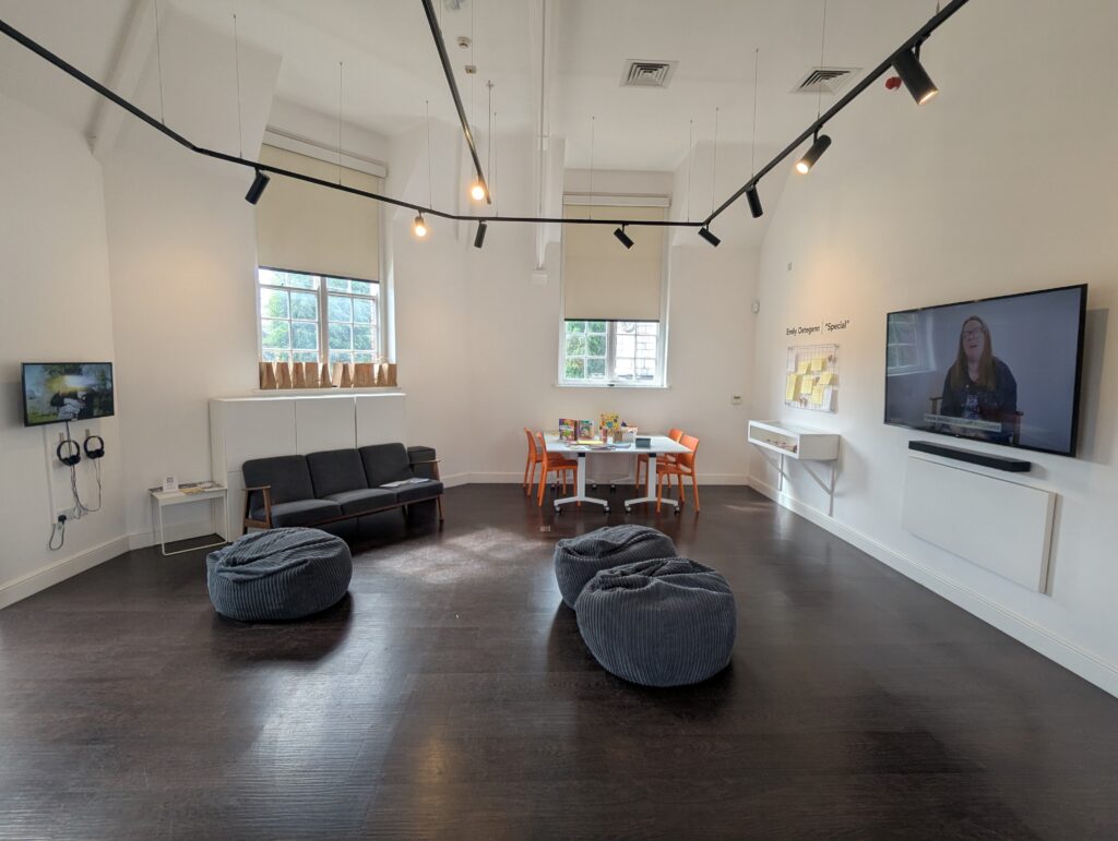 A whitewall gallery space with TVs on the left- and right-hand walls showing films, each with grey bean bags in front. On the back wall, two windows, a sofa, and an engagement table with chairs tucked beneath.