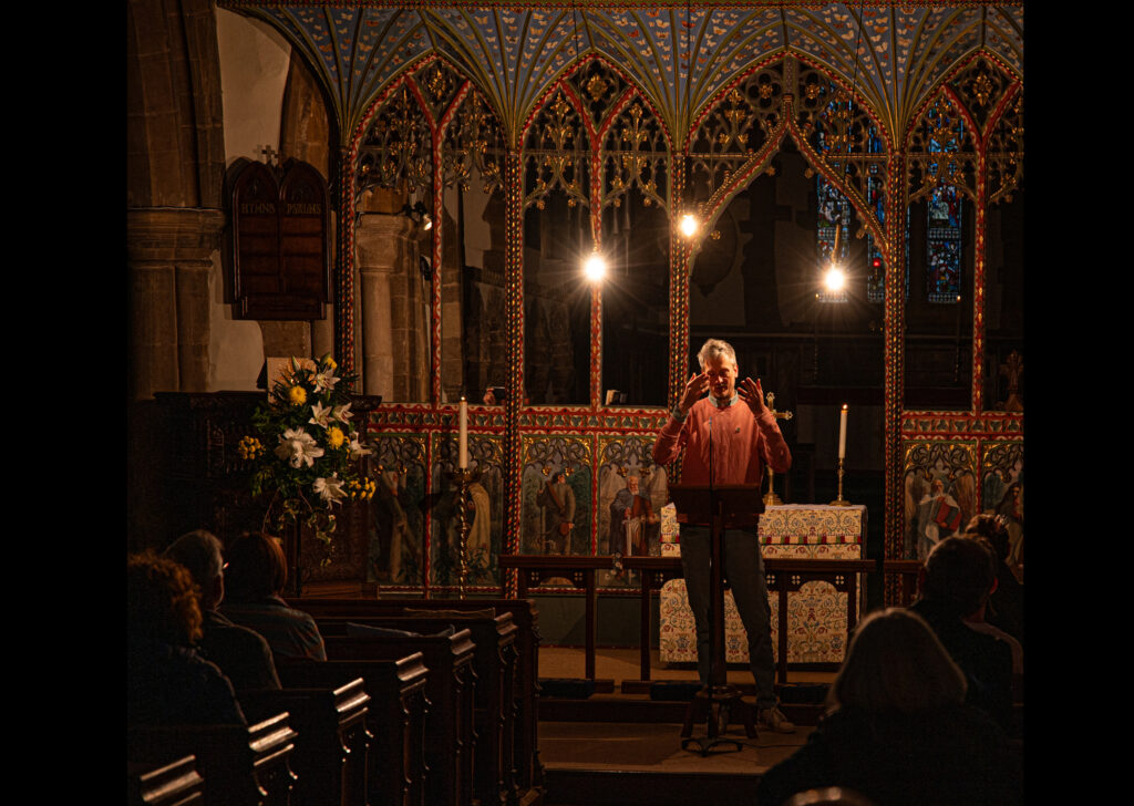 Three lights are suspended from the ceiling of a church. The lights glow warmly and illuminate the face of the speaker at the front of the church.