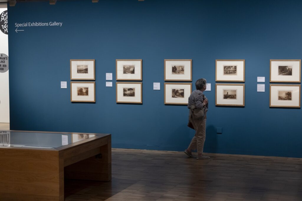 A dark blue gallery room with ten framed works hung in pairs and a viewer in front of them