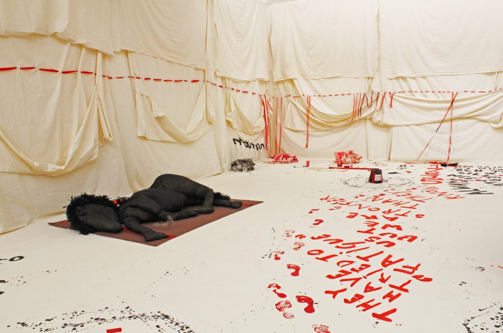 A gallery space with white vinyl flooring and walls draped with cream calico. Large red threads stitched into the calico. Large red letters painrd on the floor read They have tried to fatigue us but we are stronger than. they know.. A large stuffed fabric horse lays on a mat.