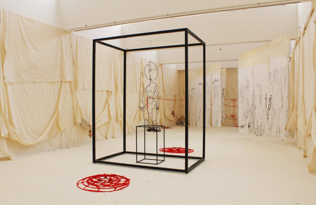 A white gallery space draped with cream coloured calico. In the centre a steel framed box shape, inside another, smaller box with a figure, made out of wire hanging above it. Two red skirt hoops on the ground on either side of the structure.