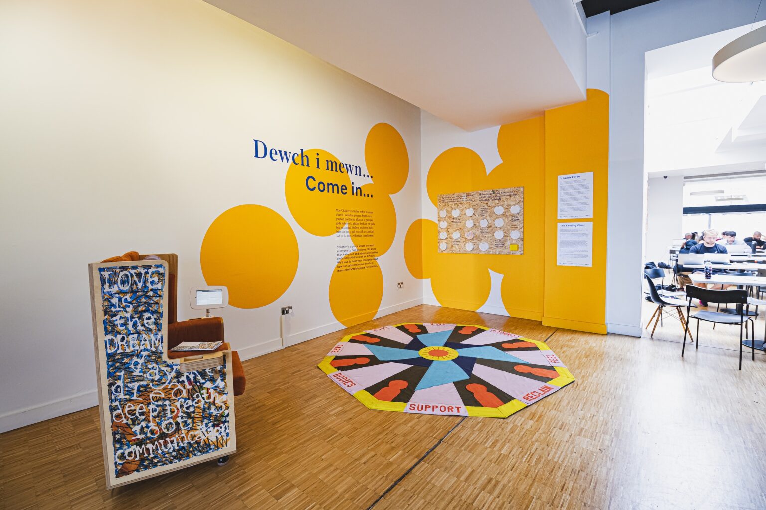 Photgraph of the corner of a brightly lit cafe corner with a large blocky chair and a hexagonal floor textile, the walls behind painted with a bright orange design with circles.