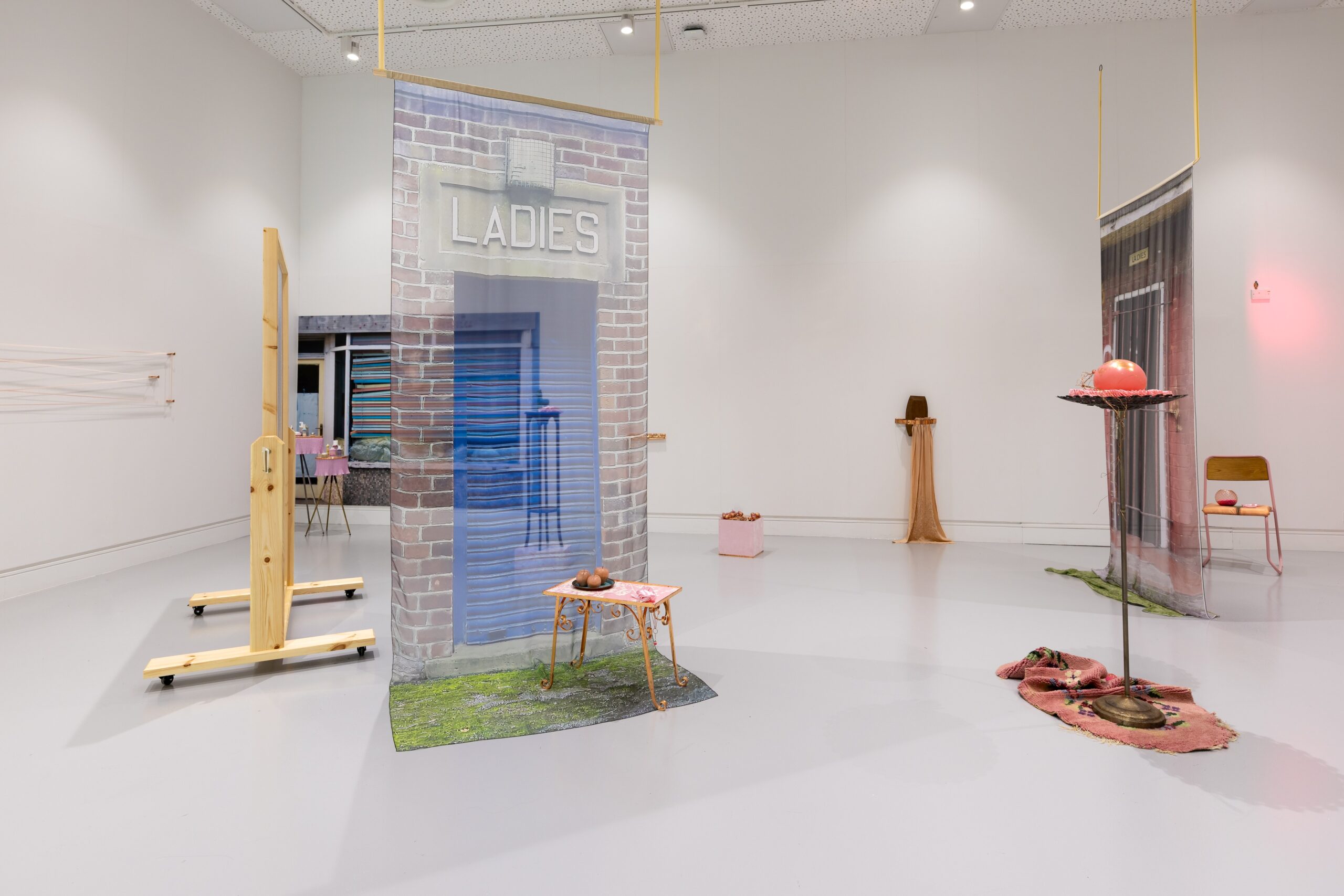 A large white wall gallery space with assemblages of objects spread around the space, chiffon prints of the doors to public bathrooms hung from the ceiling.