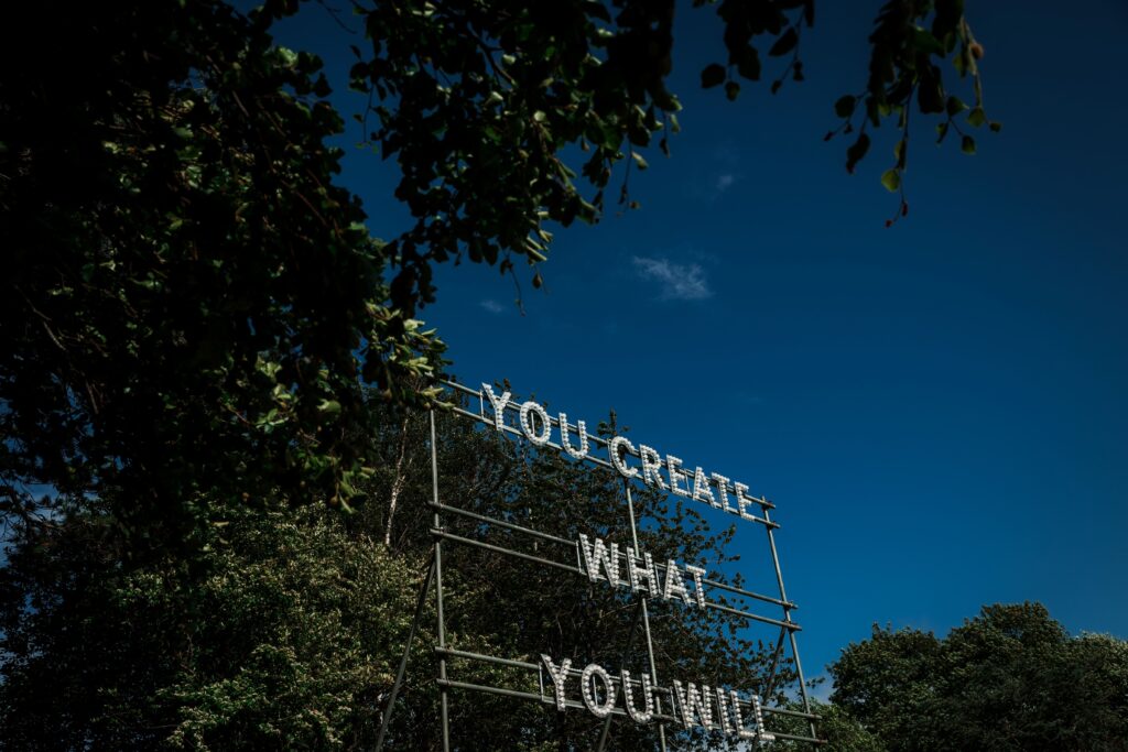 A sculptural text work set up in leafy parkland. Scaffolding supports lighting rigs, bulbs spelling out the phrase; "YOU CREATE WHAT YOU WILL".