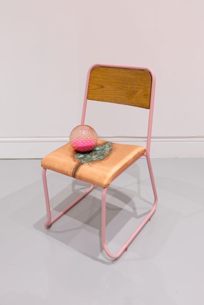 A chair with pink frame, plywood back, and satin upholstered seat printed with copper staining, a pink stippled glass ball resting on the seat..