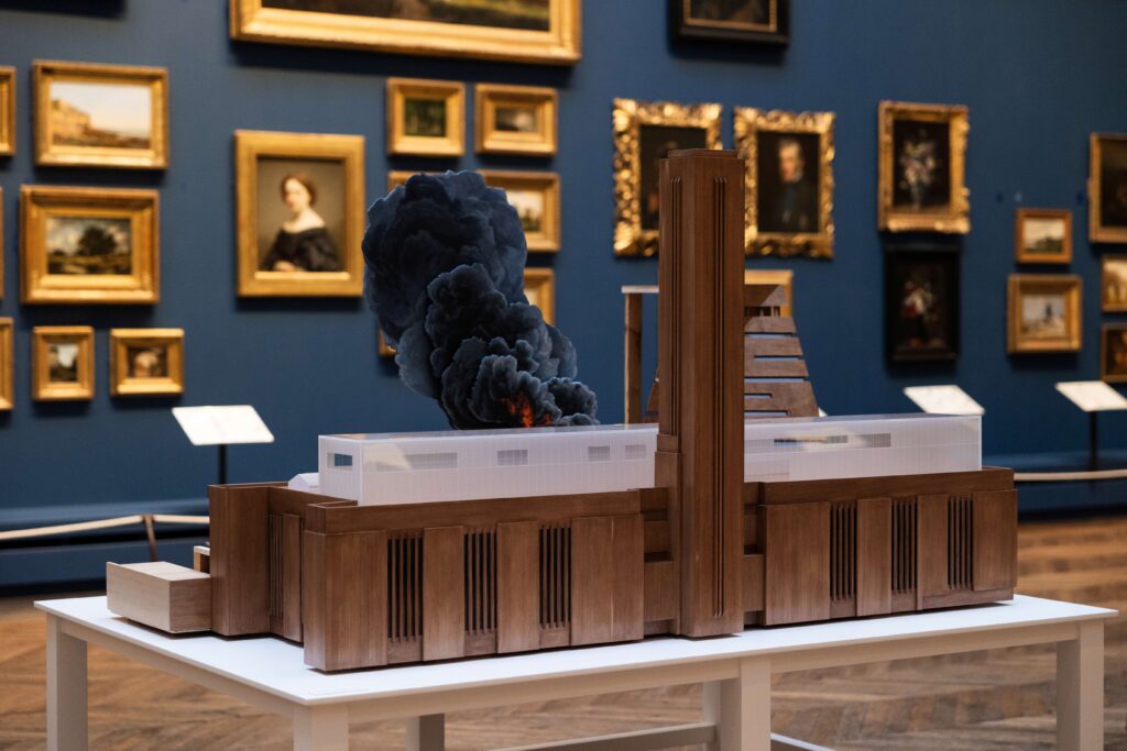 A scale model of Tate Modern on fire sits in a traditional gallery space, backed by a number of oil paintings in gold frames.