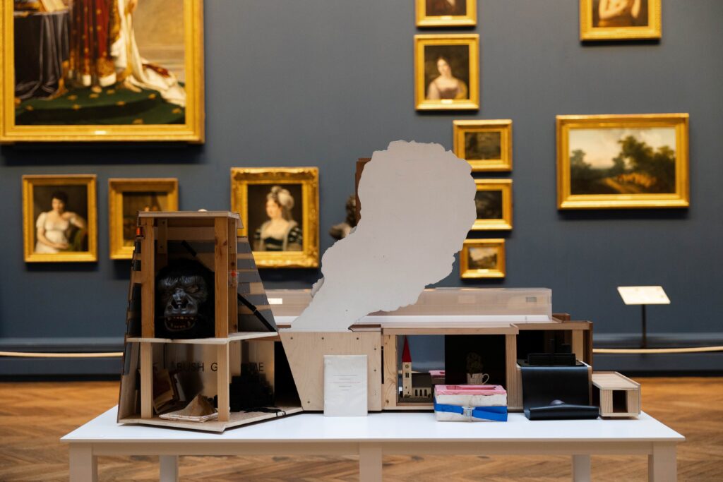 The back of a scale model of Tate Modern on fire, the cavities filled with various objects including a gorilla mask and a small painted model church. It sits in a traditional gallery space, backed by a number of oil paintings in gold frames.