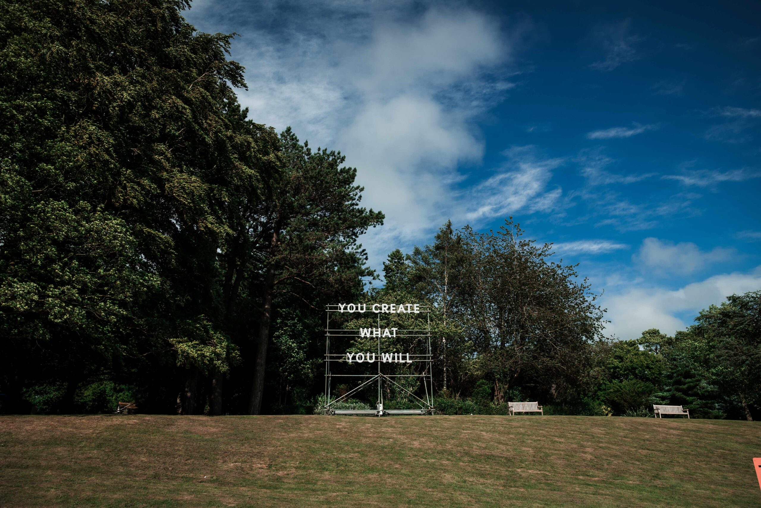 A sculptural text work set up in leafy parkland. Scaffolding supports lighting rigs, bulbs spelling out the phrase; "YOU CREATE WHAT YOU WILL".