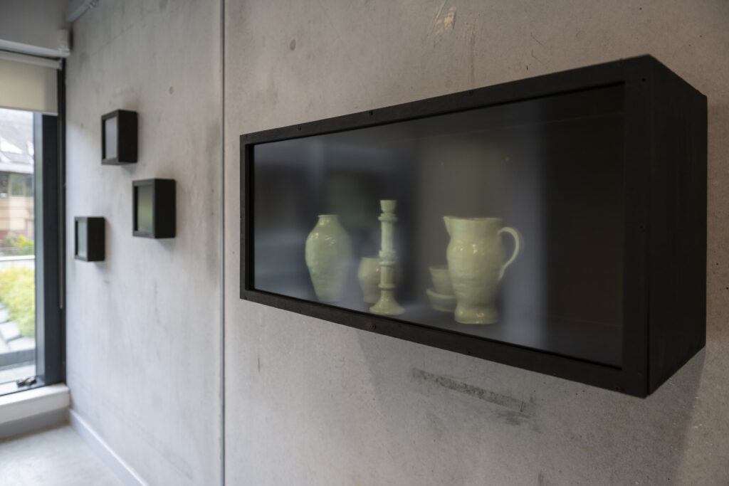 Several black wall-mounted vitrines with frosted covers on a concrete wall. The largest, closest vitrine contains several jade-coloured vessels, their outlines blurred by the frosted glass.