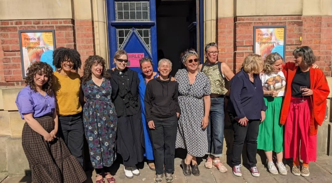A group of ten smiling women in bright clothes outside an old red brick building with a blue door ajar behind them