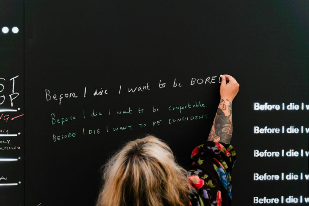 A light skinned person with long blonde hair is writing the words 'Before I die I want to be bored' on a blackboard with a piece of chalk
