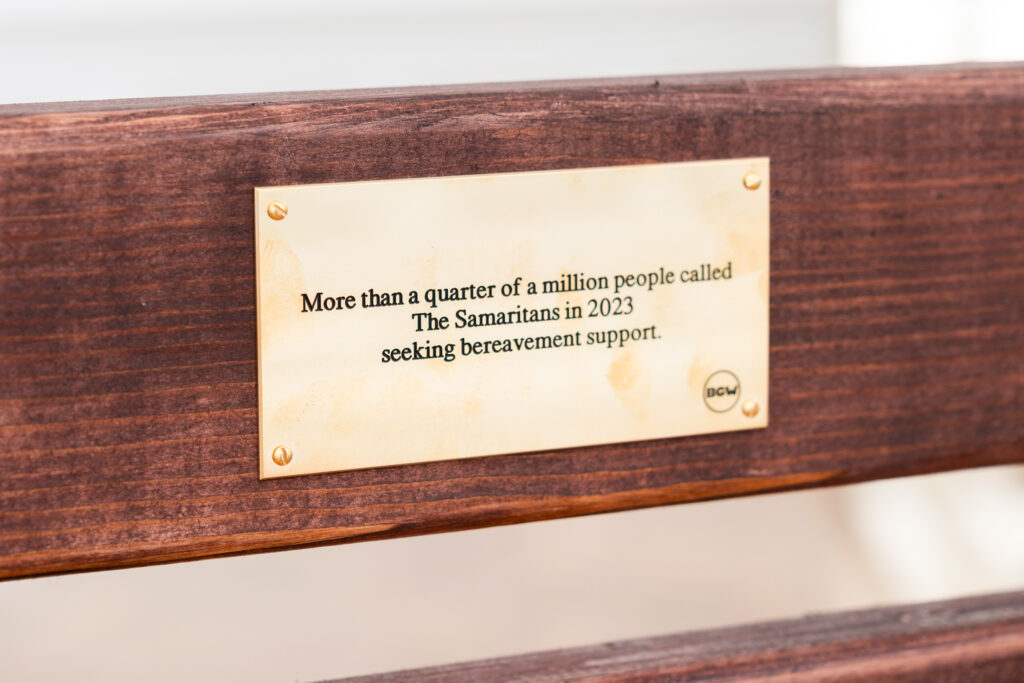 A brass plaque fixed to a wooden bench reads 'More then a quater of a million people called The Samaritans in 2023 seeking bereavement support.'